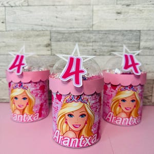 May include: Three pink party favor containers with a Barbie theme. Each container has a pink lid with a white star and a pink number 4. The containers feature a Barbie graphic and the name "Arantxa".