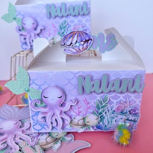 Puede incluir: Dos cajas de regalo moradas y blancas con temática de sirena. Las cajas están decoradas con un pulpo, conchas marinas y una cola de sirena. Las cajas están personalizadas con el nombre "Nalani" y el número "uno".