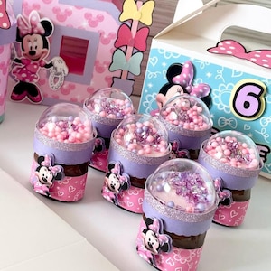 Op de afbeelding: Minnie Mouse-themafeestversieringen. De afbeelding toont kleine potjes met doorzichtige koepels gevuld met roze en paarse kralen, versierd met Minnie Mouse-afbeeldingen en glitter. Andere decoraties zijn een groot cijfer 5 en een doos met het cijfer 6.
