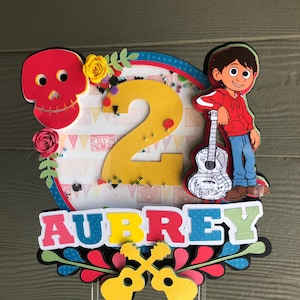 May include: A colorful cake topper featuring a cartoon character from the movie Coco, a yellow number 2, and the name Aubrey. The topper is decorated with flowers, leaves, and guitars.