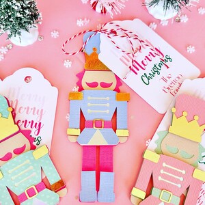 May include: Three die-cut nutcracker ornaments in pastel shades of blue, pink, and mint green with gold foil crowns and belts. Each nutcracker is attached to a white gift tag featuring the red and green text Merry Christmas and The Bretton family.