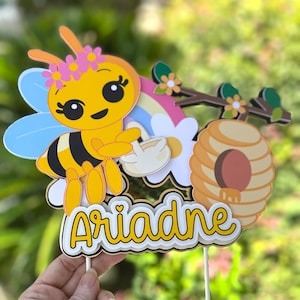 May include: A yellow and black bee-themed cake topper with the name "Ariadne" in a playful font. The topper features a cartoon bee with a flower crown, a rainbow, a beehive, and flowers. The design is held by two wooden sticks.