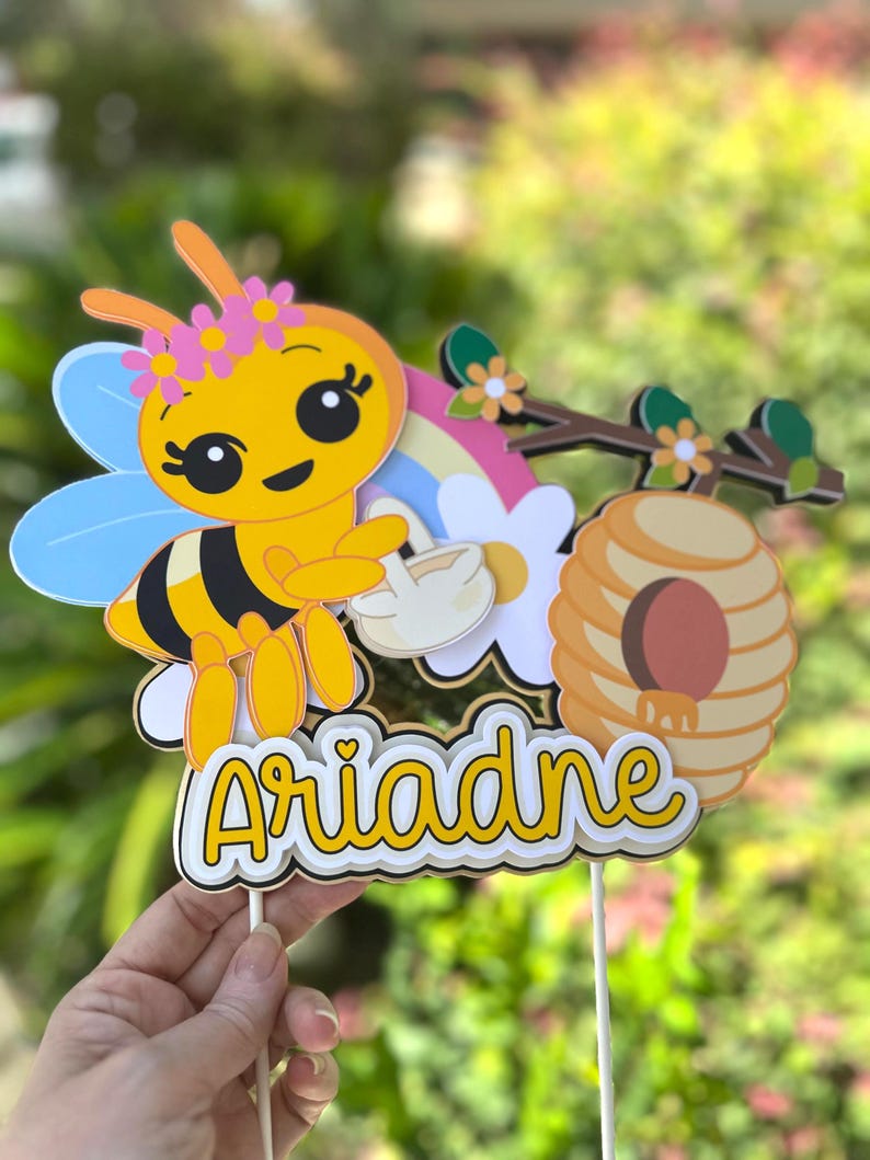 May include: A yellow and black bee-themed cake topper with the name "Ariadne" in a playful font. The topper features a cartoon bee with a flower crown, a rainbow, a beehive, and flowers. The design is held by two wooden sticks.