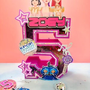 May include: A colourful, custom-made birthday cake topper shaped like the number 5, with the name "Zoey" in pink. The topper includes cartoon illustrations of three figures, stars, a blue cat, and a microphone.