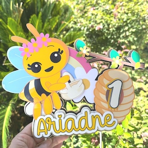 May include: A yellow and black bee-themed cake topper with the name "Ariadne" in yellow script. The topper includes a cartoon bee with a flower crown, a rainbow, a beehive with the number "1", and floral accents.