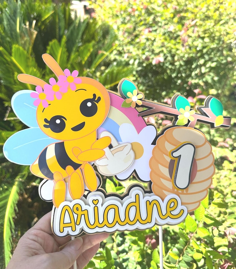 May include: A yellow and black bee-themed cake topper with the name "Ariadne" in yellow script. The topper includes a cartoon bee with a flower crown, a rainbow, a beehive with the number "1", and floral accents.