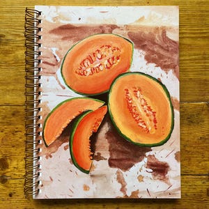 Puede incluir: Cuaderno con espiral que presenta una vibrante ilustración de melón cantalupo en rodajas. La obra de arte muestra la fruta naranja con bordes verdes y semillas visibles, sobre un fondo marrón y blanco. La encuadernación en espiral del cuaderno es dorada.