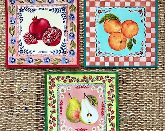 Puede incluir: Tres baldosas decorativas cuadradas con ilustraciones de frutas. Una baldosa presenta una granada, otra naranjas y la tercera peras. Cada baldosa tiene un borde decorativo con motivos florales y de hojas. Las baldosas están sobre una superficie tejida.