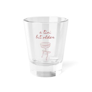 May include: A clear shot glass with the phrase "a tini bit older" in red script. Below the text is a red illustration of a cocktail glass with a bow. The glass has a thick base and straight sides.