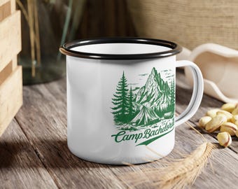 May include: White enamel camping mug with a black rim. The mug features a green illustration of a mountain range, trees, a tent, and the text "Camp Bachelor".