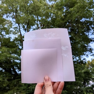 May include: Three sheets of light purple, perforated fabric are held up against a backdrop of green trees and blue sky. The fabric has a fine, dotted texture and appears to be semi-transparent. The sheets are different sizes, with the largest at the top.