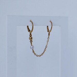 Peut inclure: Une boucle d'oreille double anneau dorée reliée par une fine chaîne. Un anneau est orné d'une petite perle dorée et d'une perle blanche suspendues, tandis que l'autre anneau est simple. Le bijou présente une finition polie et un fermoir sécurisé.
