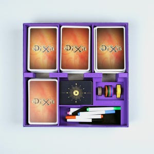 May include: Purple game box with a Dixit logo on the front. The box contains a set of cards with colorful illustrations, a score pad, and markers.