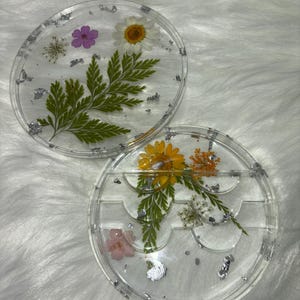May include: Two clear round resin trays with dried flowers and silver leaf accents. The top tray has a fern, a white flower, and a purple flower. The bottom tray has a sunflower, a white flower, and a pink flower. The bottom tray has three sections for holding lashes.