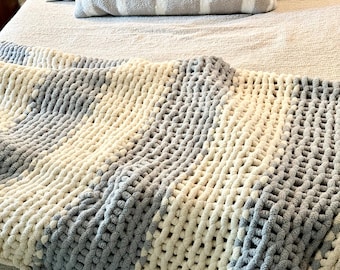 May include: A chunky knit throw blanket in a light gray and cream color pattern. The blanket is made of soft, fluffy yarn and has a textured, waffle-like weave.