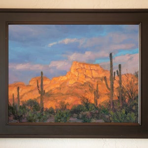 May include: A framed oil painting depicting a desert landscape with tall cacti in the foreground and a large, rocky mountain in the background. The sky is a vibrant blue with fluffy white clouds. The painting is framed in a dark brown wood frame.