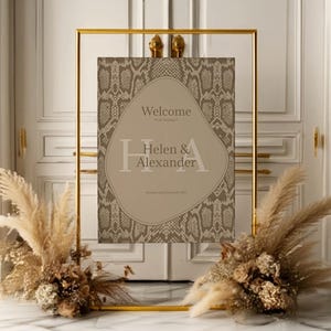 May include: A gold metal framed welcome sign with a snakeskin pattern background. The sign reads "Welcome Helen & Alexander" in a soft, neutral color. Dried floral arrangements flank the sign.