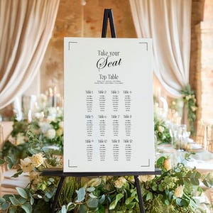 Op de afbeelding: Een witte trouwzittingstabel op een zwart schildersezel, met de woorden "Take your Seat" en "Top Table" in elegant schrift. De tabel geeft de tafelnummers en namen weer. De scène is versierd met bloemen en groen.