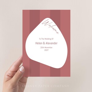 May include: A welcome sign with a white, abstract shape on a pink and white striped background. The text reads "Welcome to the Wedding of Helen & Alexander 20th November 2027".