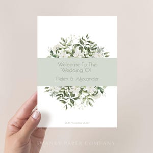 Könnte beinhalten: Eine weiße Hochzeitseinladung mit floralen Akzenten. Die Einladung hat ein hellgrünes Band mit dem Text "Welcome To The Wedding Of Helen & Alexander". Das Datum "20th November 2021" ist unten aufgedruckt. Das Logo der Swanky Paper Company befindet sich unten.