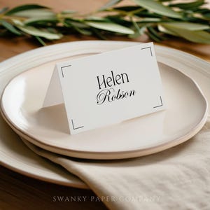 以下が含まれることがあります： エレガントな黒い筆記体で「Helen Robson」と書かれた、折り畳まれた白い席札。クリーム色の皿の上に置かれ、リネンのナプキンと背景に緑があります。