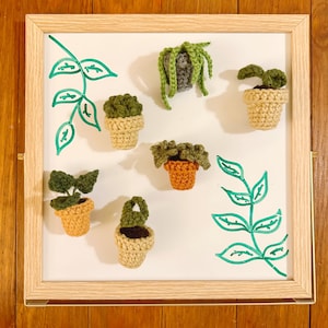 May include: A white board with a green marker drawing of a plant and seven crocheted potted plants. The plants are in various shades of green and brown.