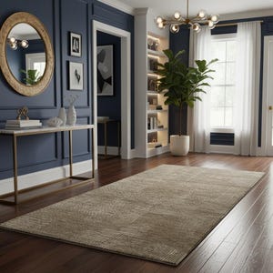 May include: A beige rectangular area rug with a geometric pattern lies on a dark wood floor. The rug is in a room with navy blue walls, a gold-framed mirror, and a console table. A large potted plant and a chandelier are also visible.