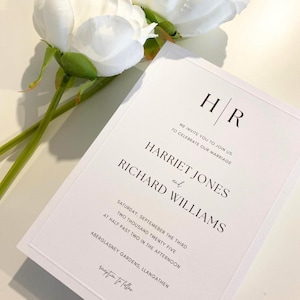 May include: A formal white wedding invitation with black text, including the names Harriet Jones and Richard Williams. The invitation details the date, time, and location. White flowers are visible in the background.