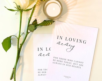 May include: Two white cards with black text that reads "In loving memory" and "For those who cannot be here today, but are always in our hearts". The cards are on a white surface with a white rose and a glass candle holder.