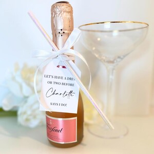 Może przedstawiać: Mała butelka szampana z białą wstążką i słomką. Biała etykieta jest przymocowana do butelki z tekstem "Let's have a drink or two before Charlotte says I do!"