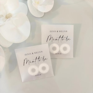 May include: Two small, square, translucent packets containing white mints. The packets are printed with the text "OLIVIA & WILLIAM" above "Mint to be" in cursive, and the date "22.09.26". White flower petals are in the background.
