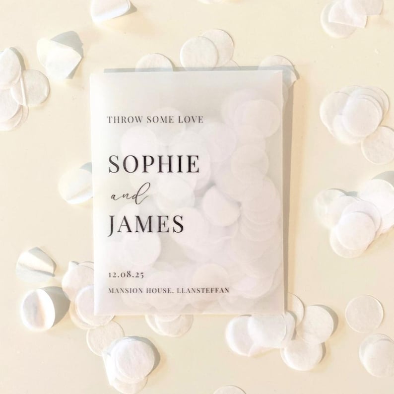 Op de afbeelding: Een transparant zakje gevuld met witte confetti, met de tekst "THROW SOME LOVE", de namen "SOPHIE and JAMES", de datum "12.08.25" en "MANSION HOUSE, LLANSTEFFAN" in zwart. Witte confetti is rondom het zakje verspreid.