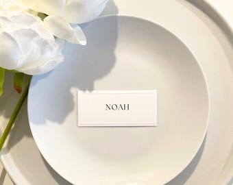 Puede incluir: Una mesa blanca con un plato y un plato más pequeño encima. Una tarjeta de sitio con el nombre "NOAH" está en el plato. Flores blancas en la esquina superior izquierda.