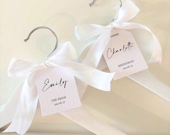 May include: Two white wooden hangers with white satin ribbon bows and personalized tags. The tags are white with black text. One tag says "Emily The Bride 09.06.25" and the other tag says "Charlotte Bridesmaid 09.06.25".