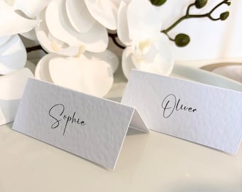 May include: Two white place cards with textured paper, each folded in half. One card reads "Sophie" in elegant black script, the other reads "Oliver." White orchids and greenery are in the background, creating a soft, elegant aesthetic.