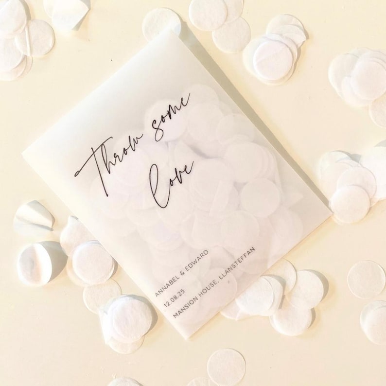 Op de afbeelding: Een doorschijnend wit zakje gevuld met witte confetti, verspreid op een lichtgele ondergrond. Op het zakje staat de tekst "Throw some Love" in sierlijke letters. Namen en een datum zijn ook gedrukt.