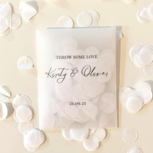 Op de afbeelding: Een doorschijnend zakje gevuld met witte confetti, met de tekst "THROW SOME LOVE" en de namen "Kirsty & Oliver" in sierlijke letters. De datum "21.09.25" staat ook op het zakje. Witte confetti is rond het zakje verspreid.