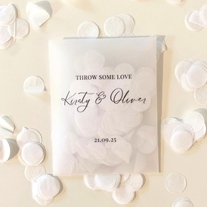 Op de afbeelding: Een doorschijnend zakje gevuld met witte confetti, met de tekst "THROW SOME LOVE" en de namen "Kirsty & Oliver" in sierlijke letters. De datum "21.09.25" staat ook op het zakje. Witte confetti is rond het zakje verspreid.
