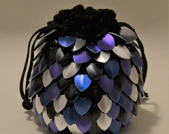 May include: A round, scale-covered pouch in shades of black, silver, and purple. The pouch has a drawstring closure and a black, textured top. The scales are arranged in an overlapping pattern, creating a textured surface.