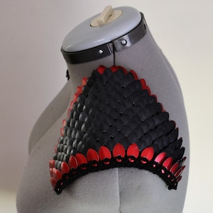 May include: A black and red scale mail shoulder piece. The scales are made of metal and are arranged in a pattern. The shoulder piece is on a gray mannequin.