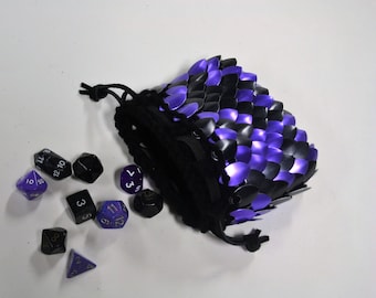 May include: A black drawstring dice bag with a scale mail design in black and purple. Several polyhedral dice in black and purple are scattered around the bag, including D4, D6, D8, D10, D12, and D20.