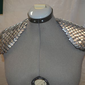 Puede incluir: Armadura de hombro de cota de malla plateada en un maniquí. La armadura está hecha de pequeñas escamas de metal superpuestas.