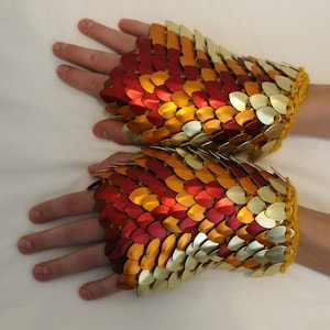 May include: A pair of fingerless gloves made of metal scales in shades of gold, red, and orange. The gloves are crocheted with a yellow yarn at the wrist.