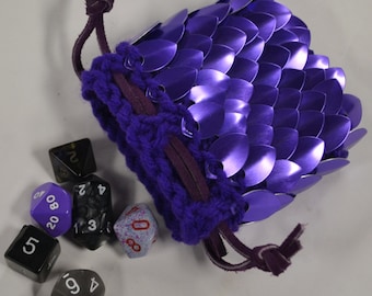 May include: A purple scale mail dice bag with a drawstring closure. The bag is made of overlapping metal scales and has a knitted purple top. Several multi-sided dice in various colors are scattered around the bag.