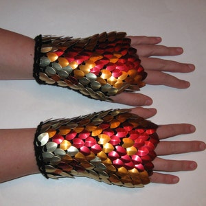 Puede incluir: Un par de guantes de malla de cadena roja, dorada y plateada. Los guantes están hechos de pequeñas escamas de metal superpuestas.
