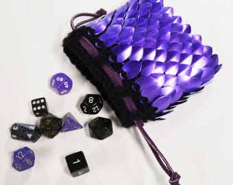 May include: A purple scale mail dice bag with a black trim and drawstring. Several multi-sided dice in various colors and shapes are scattered around the bag, including a black six-sided die with white dots and a purple twenty-sided die.