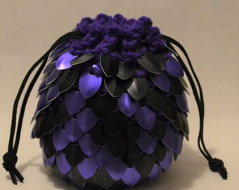 May include: A round, dragon-scale inspired pouch in black and purple. The pouch is made of overlapping metal scales, with a drawstring closure made of purple yarn and black cord. The pouch is designed to resemble a dragon egg.