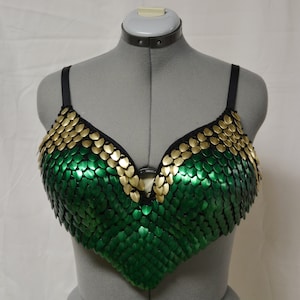 May include: A green and gold bra made of metal scales. The bra has a V-shaped neckline and adjustable straps.