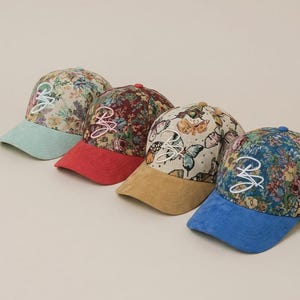 Peut inclure: Quatre casquettes de baseball avec des motifs floraux et de papillons. Chaque casquette a une visière de couleur différente : bleu clair, rouge, beige et bleu. Les casquettes ont un logo "B" brodé en blanc.