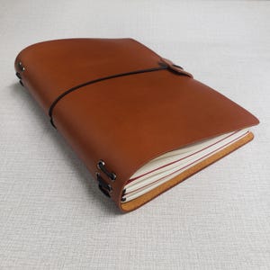 May include: A brown leather journal with a black elastic closure. The journal has a stitched binding and a strap closure. The pages are white with red lines. The journal is closed and lying on a light gray surface.